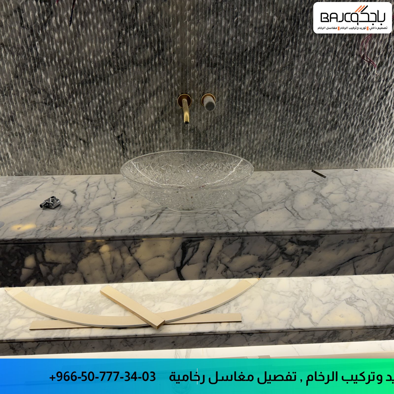 مغسلة رخام أبيض مع عروق رمادية وحوض زجاجي شفاف، وجدار بلاط رمادي منقوش، وخلاطات ذهبية