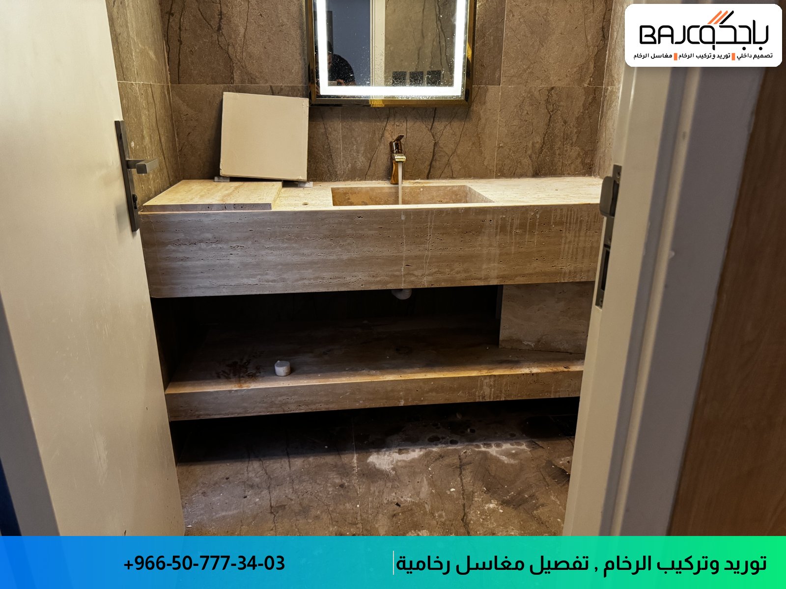 مغسلة رخام حائطية بحوض واحد وسطح بني داكن بعروق طبيعية، مع دولاب خشبي تحتها وخلاط فضي، ومرآة مضاءة أعلاها