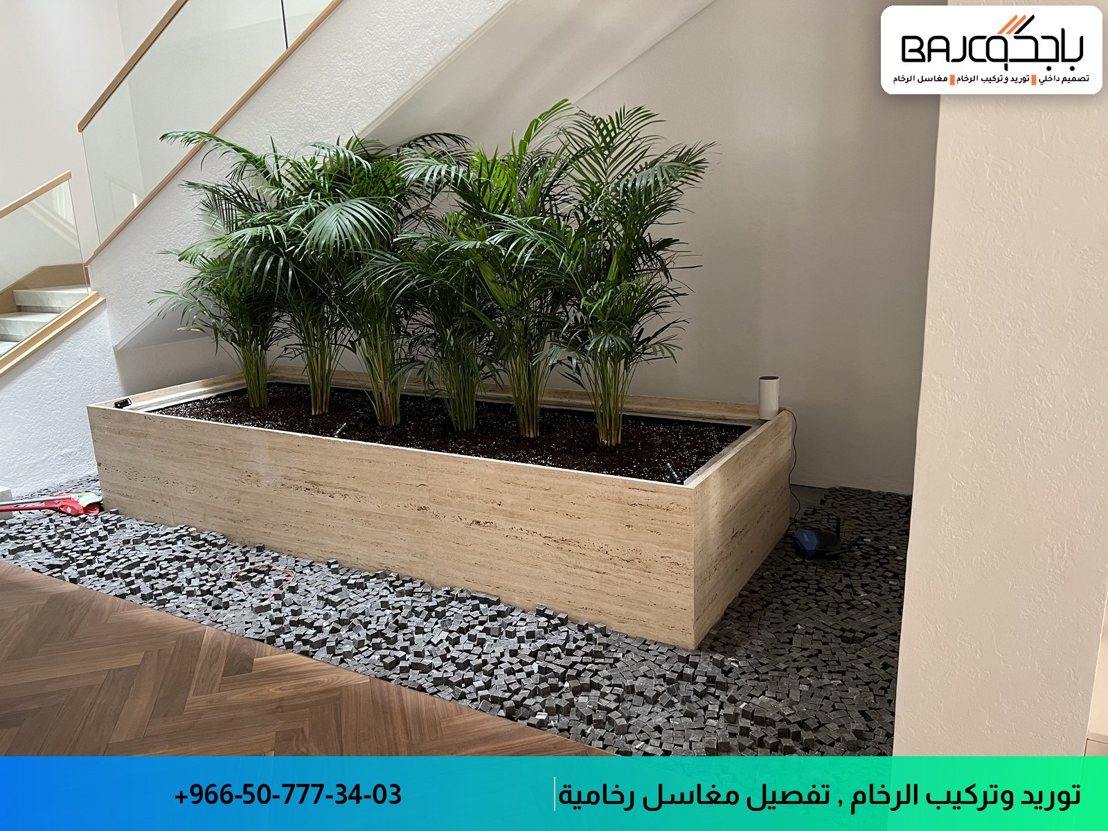 حوض رخام كبير بإطار خشبي فاتح يحتوي على نباتات خضراء، مثبت في مدخل بأرضيات حصى رمادية وبلاط باركية